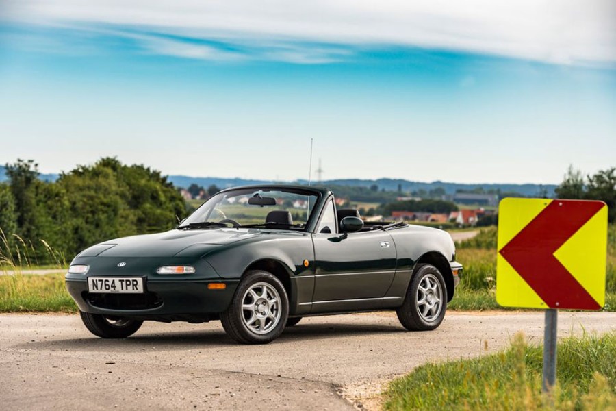 Front left shot of a dark green Mazda MX-5 parked next to red and yellow sign