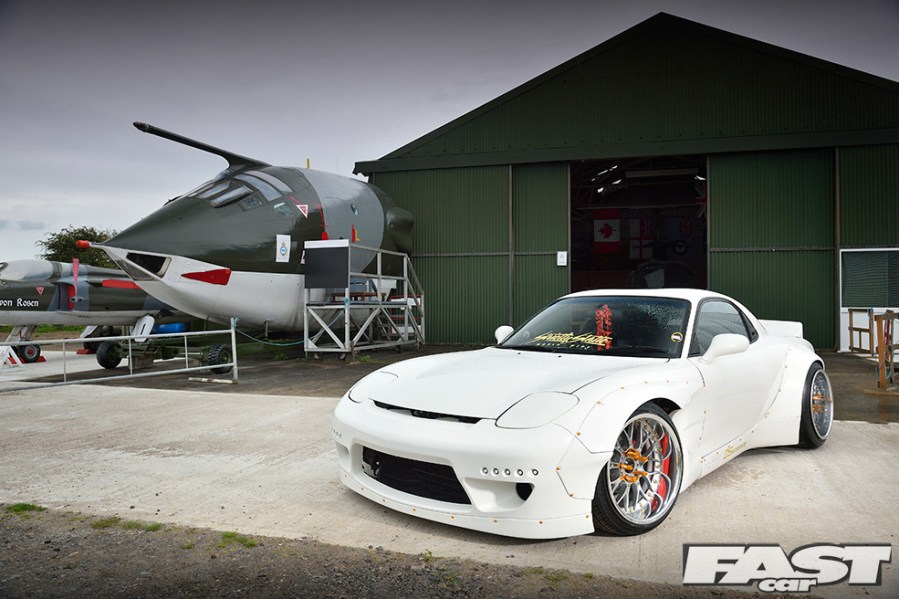 A front left side shot of a white Mazda RX7 next to an aircraft and storage garage