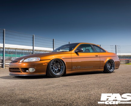 A left side view of an orange Lexus Soarer Z30 parked in front of a wire fence
