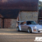 A stone brick building behind a right side shot of a silver Porsche 964