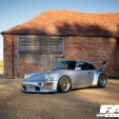 The left side of a silver Porsche 964 with a sunlit red brick building behind