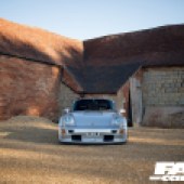 A distanced central shot of a silver Porsche 964 with a sunlit red brick building behind