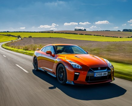 A front right shot of a bright orange Nissan GT R R35 driving through bright green fields