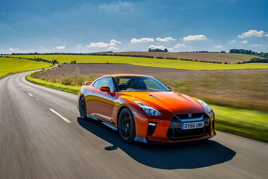 A front right shot of a bright orange Nissan GT R R35 driving through bright green fields