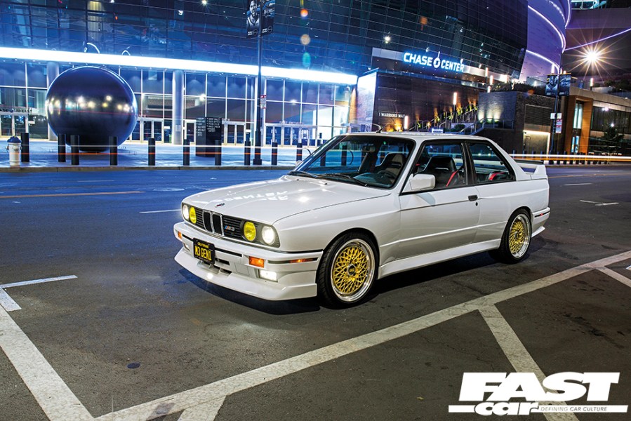 A front left side shot of a white BMW E30 M3 with gold detailing at night