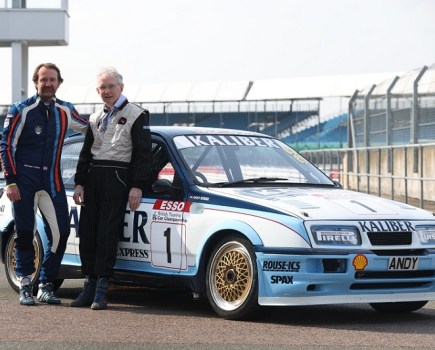 Two men leaning against the right side of a Rouse Ford Sierra RS500 on a racetrack