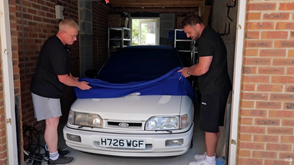 Two men peel back car cover to reveal white Ford Sierra Sapphire Cosworth parked in a garage