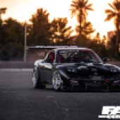 Front shot of a black Rotor Mazda RX 7 with an orange sunset and palm trees behind