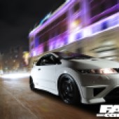 A close up front right shot of white Honda Civic FN2 TYPE-R TURBO 00 driving in city with blurred lights behind
