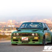 A frontal shot of a green LS-swapped BMW E30 with a city landscape behind