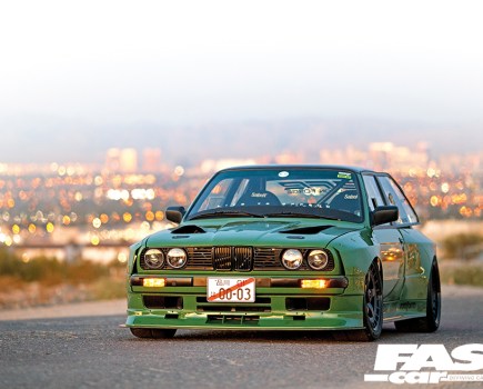 A frontal shot of a green LS-swapped BMW E30 with a city landscape behind