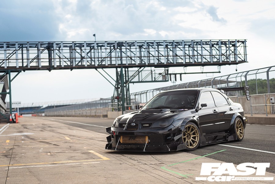 Front left shot of a black Hawkeye Impreza parked on an empty racetrack with gold alloys