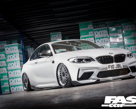 A low right side front shot of a white BMW M2 with a wall of green boxes behind