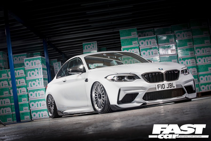 A low right side front shot of a white BMW M2 with a wall of green boxes behind