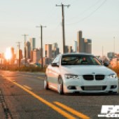 A front right view of a white tuned BMW E92 335i with a city landscape behind