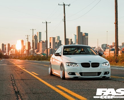 A close right front shot of a white tuned BMW E92 335i with golden sun and a city landscape behind
